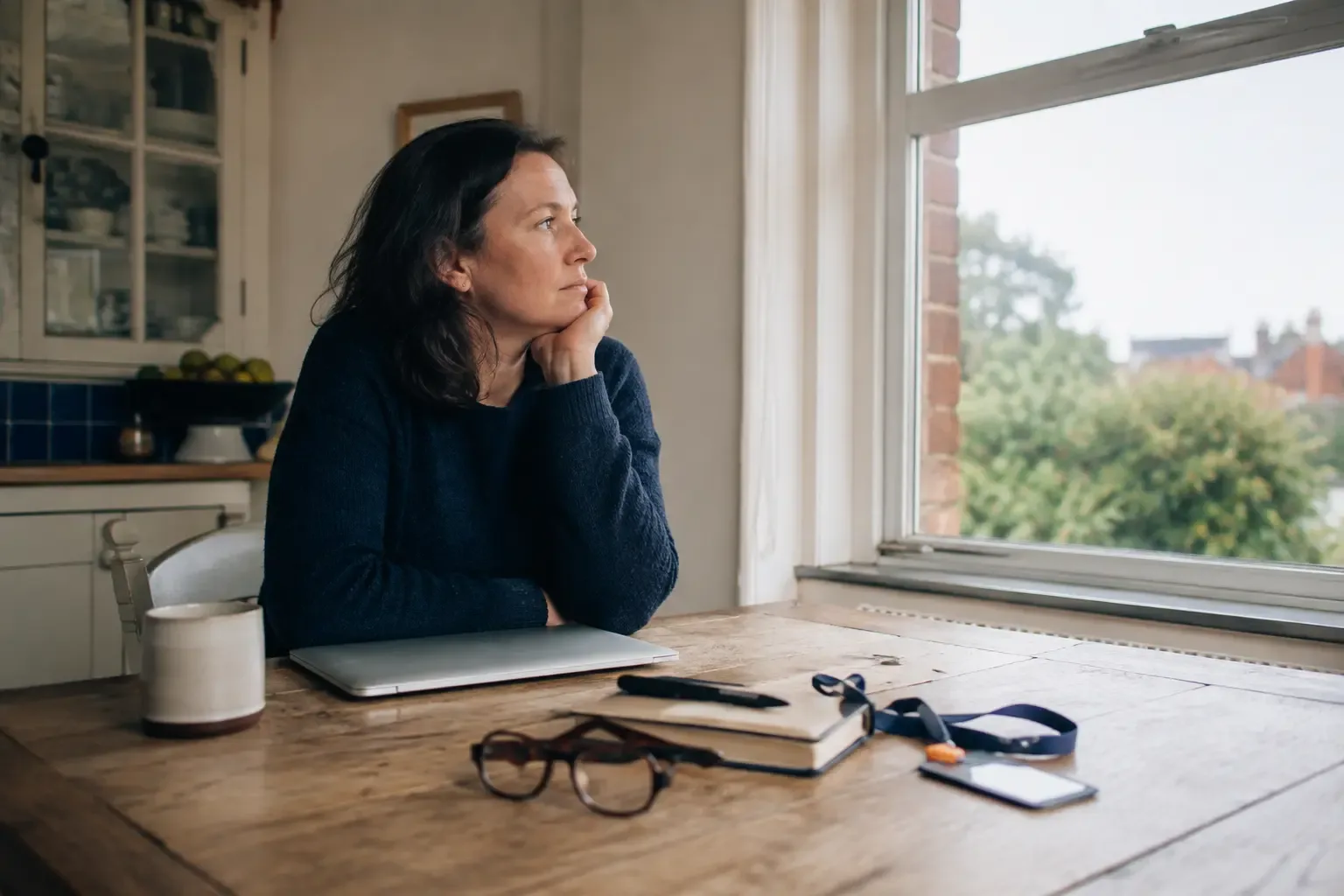 A woman pausing at a kitchen table beside her closed work laptop.