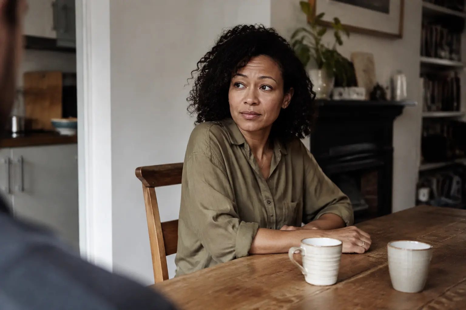 A woman sitting across a table from someone, two mugs between them, deciding what to say
