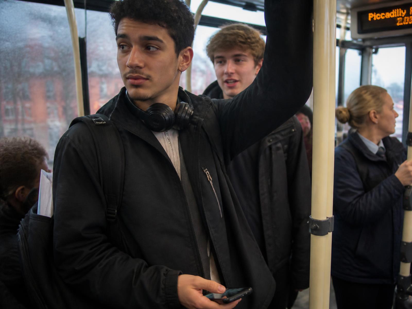A young man standing on a crowded city bus at rush hour, headphones round his neck and a phone held face down, glancing at something outside the frame while a friend talks behind him