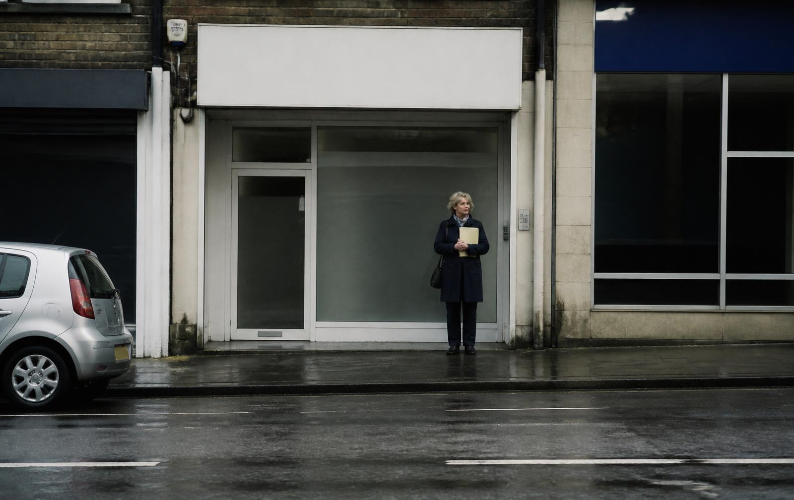 A woman of about sixty standing in a workplace corridor outside a closed office door