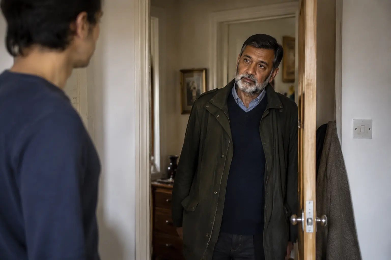 An older man standing in the doorway of his adult child's home, listening rather than speaking
