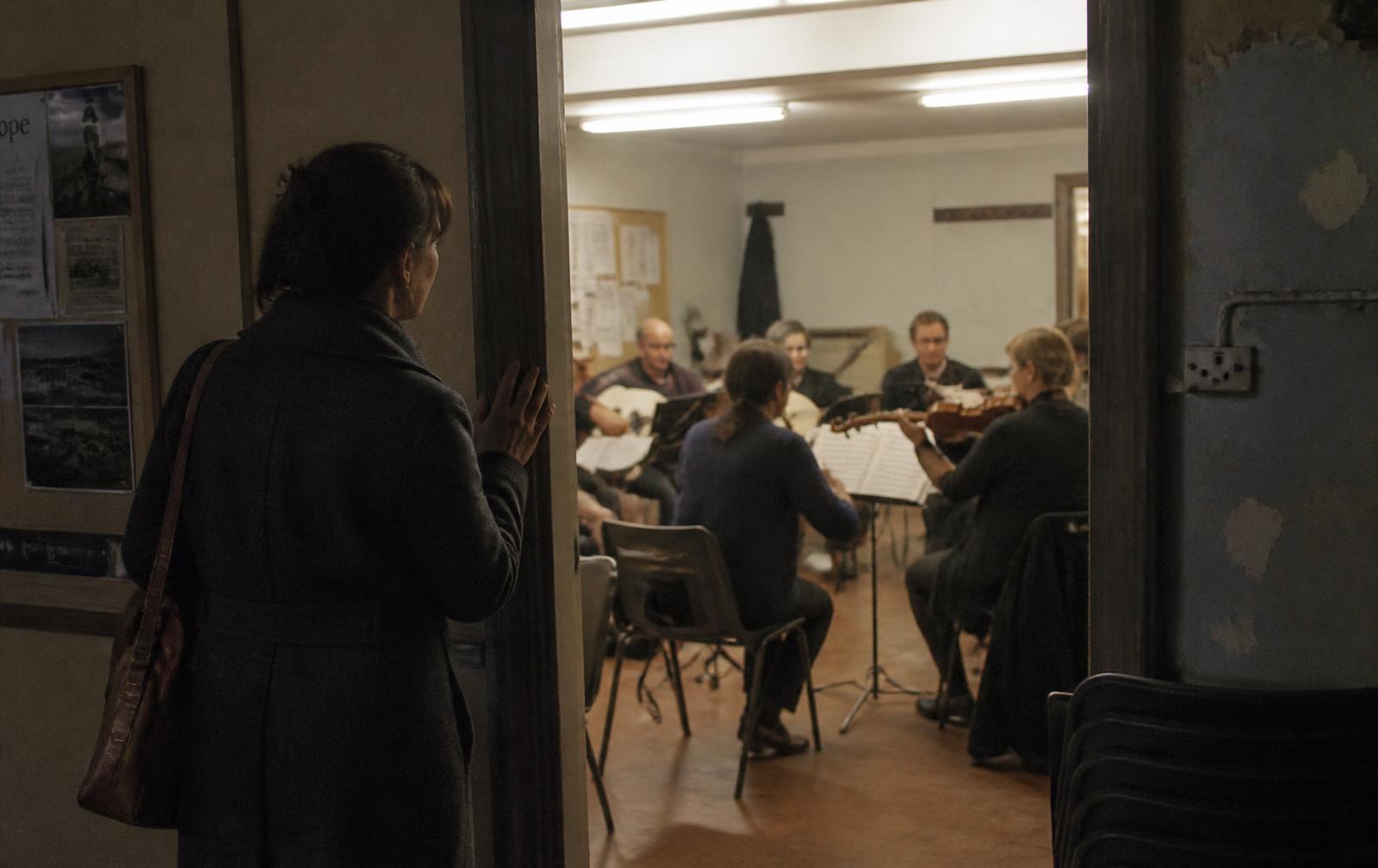 A woman standing at the side of a rehearsal she is not part of, coat still on