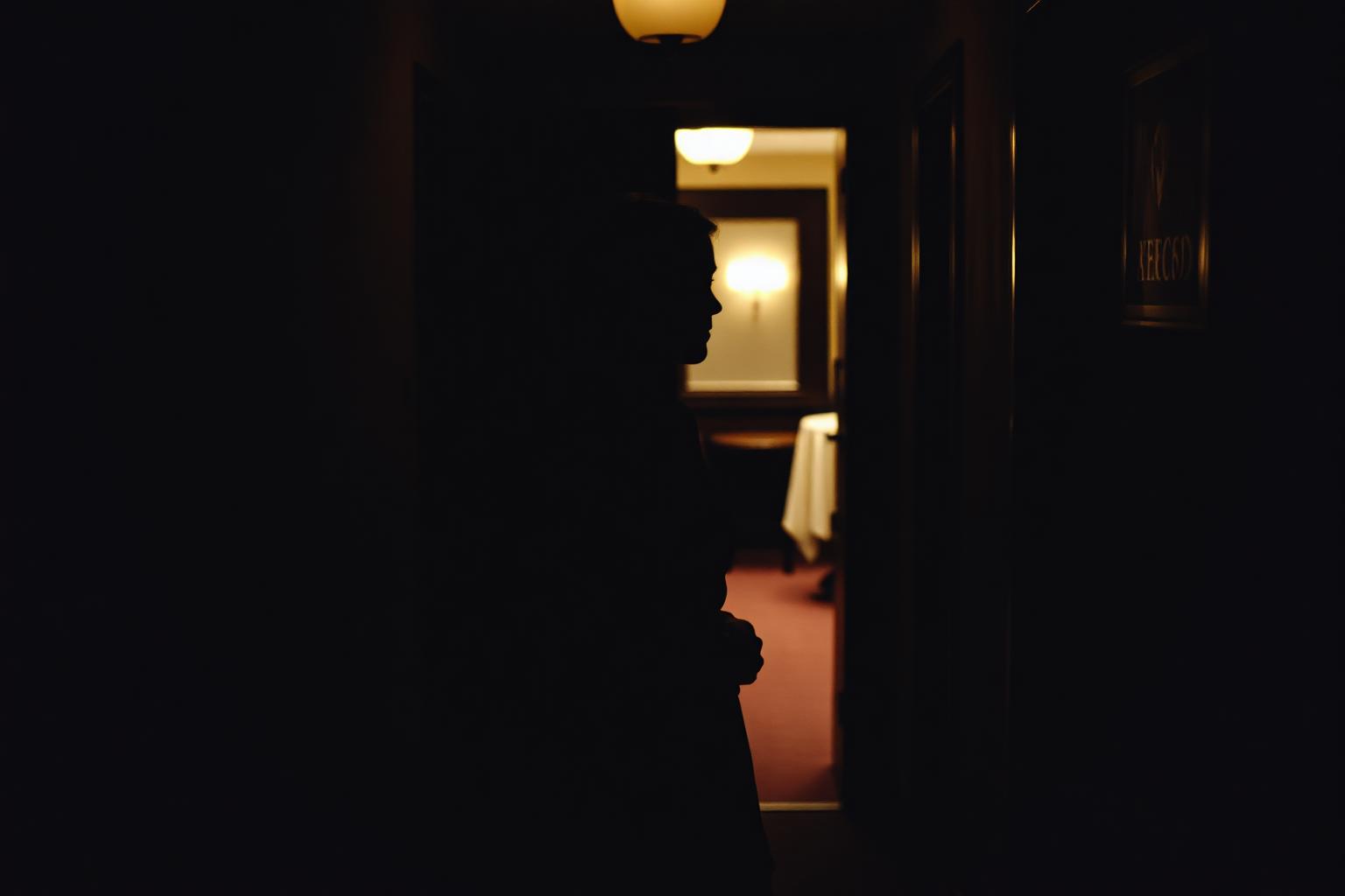 A woman in her late twenties in a dark corridor outside a lit private dining room, coat over her arm