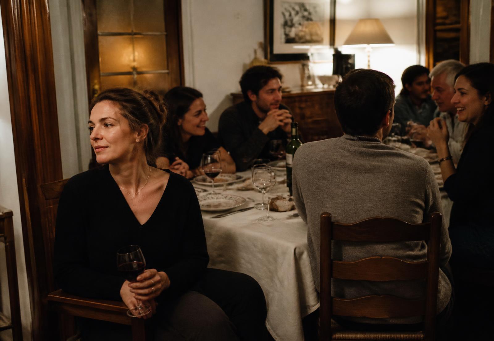 A woman sitting slightly back from a busy family dinner table, holding a glass, her attention elsewhere