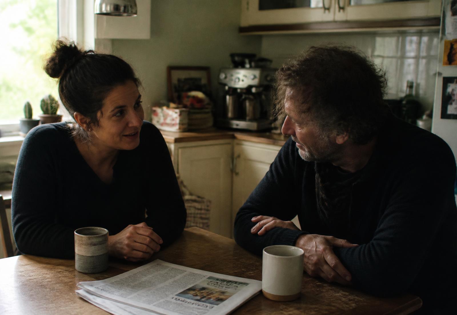 Two people at a kitchen table in the morning, one speaking and the other listening closely over mugs of tea