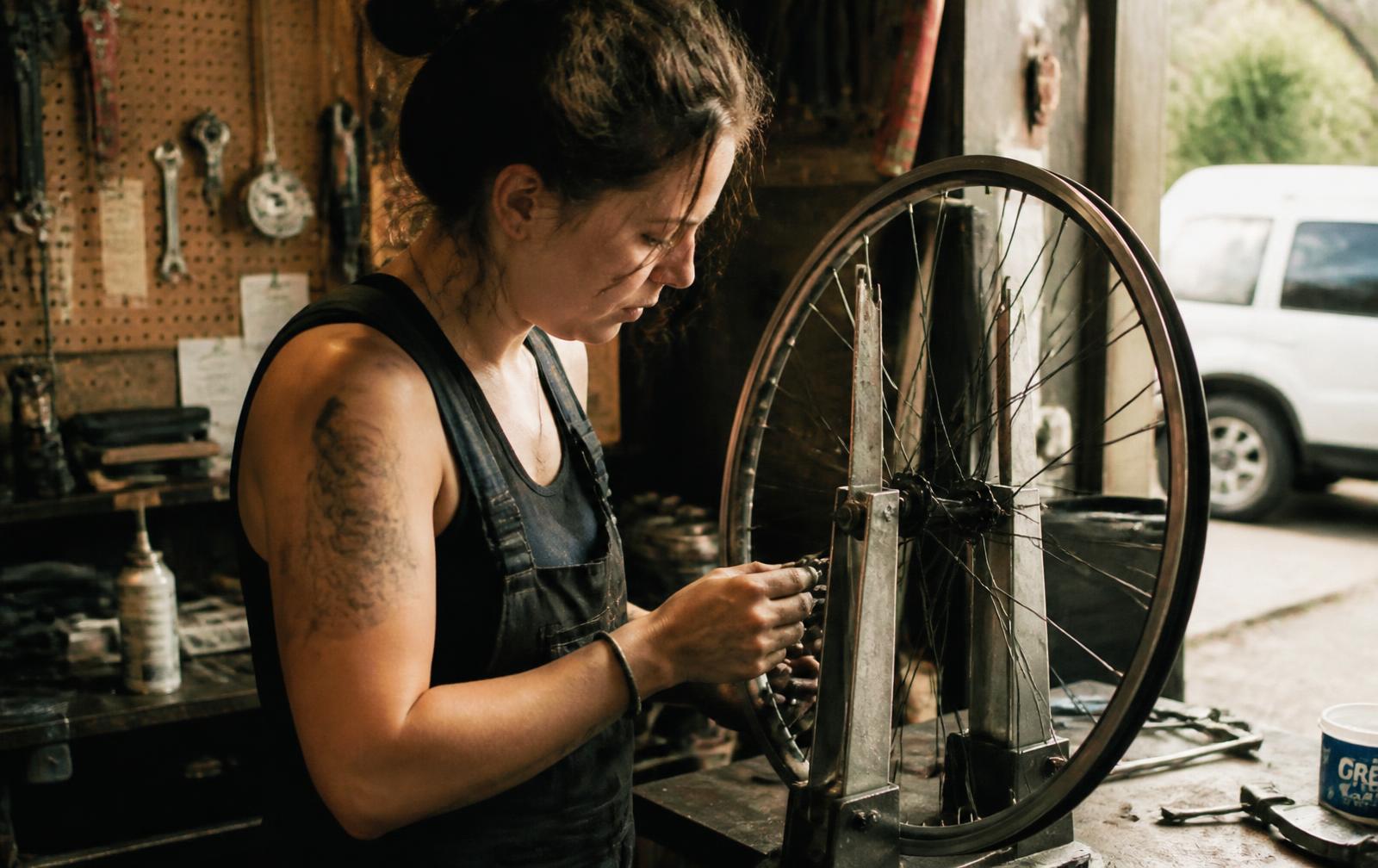 A young woman truing a bicycle wheel in a cluttered garage, absorbed in the work