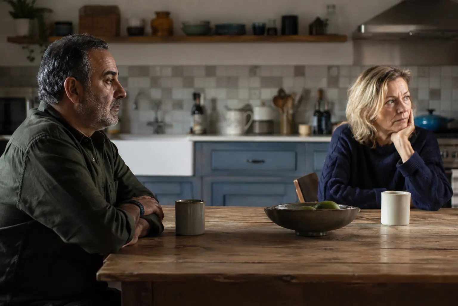 Two people sitting at either end of a kitchen table, both looking away, mid-disagreement