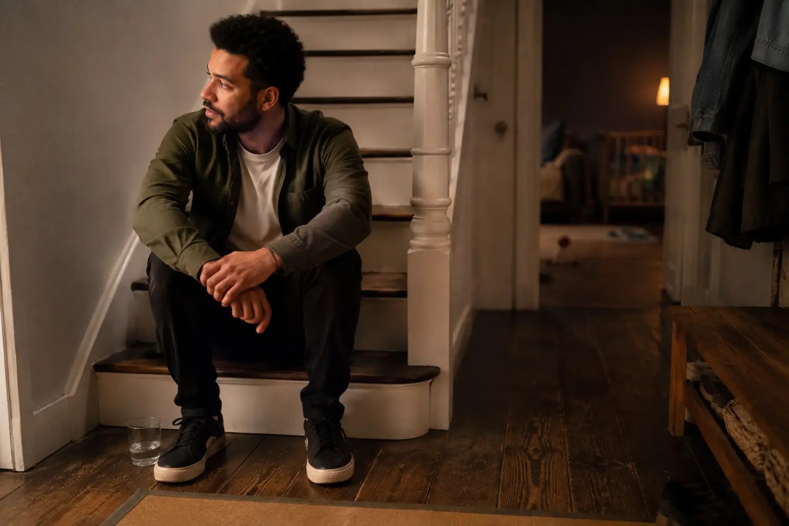 A father sitting on the stairs in a quiet hallway after putting a child to bed, a glass of water beside him