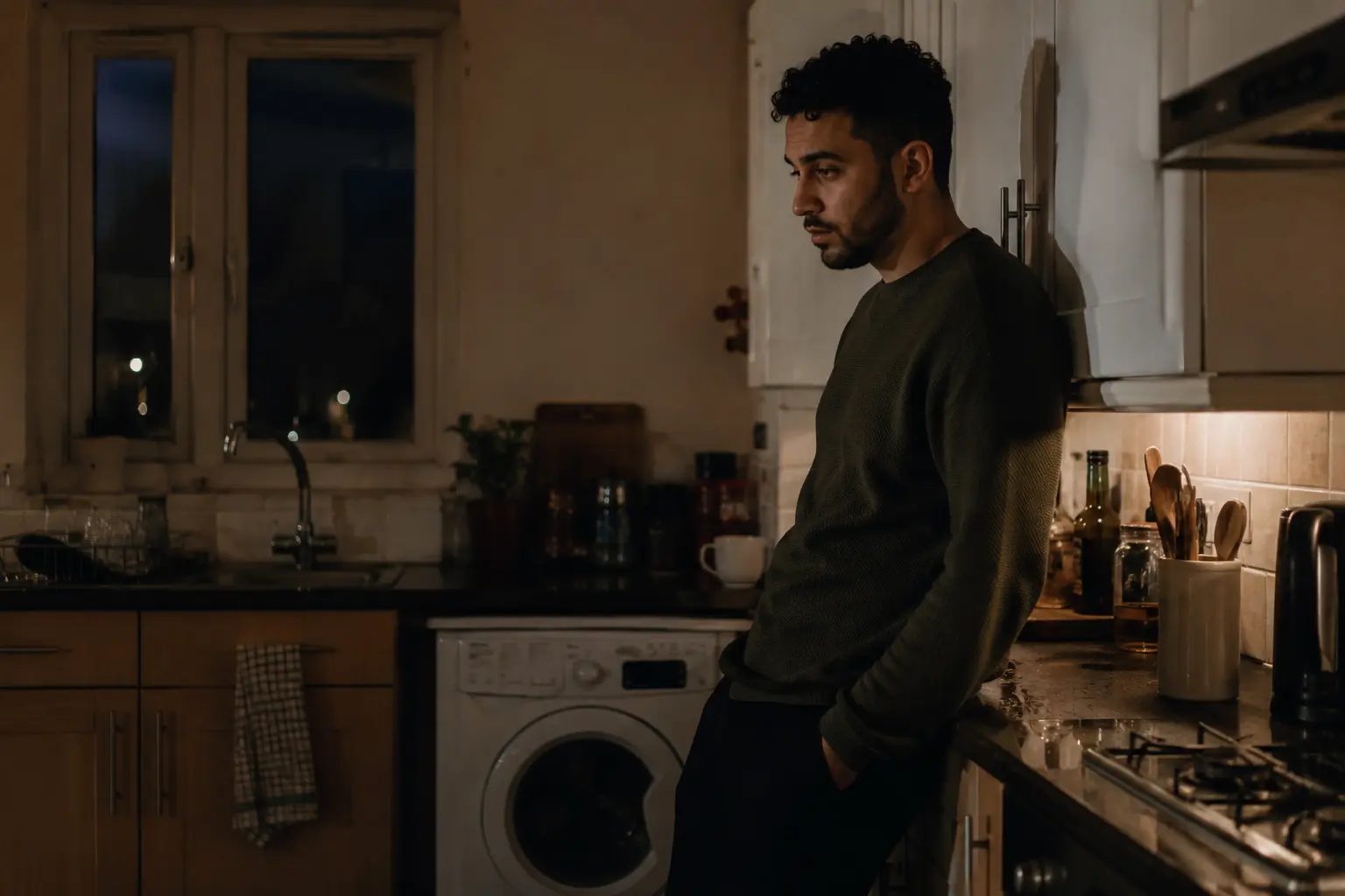 A young man paused against the counter of a dim kitchen late at night, mid-decision