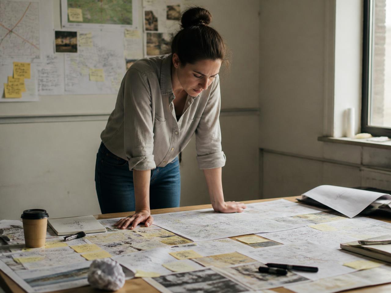 A woman standing over a table covered in maps, plans and notes, working something out